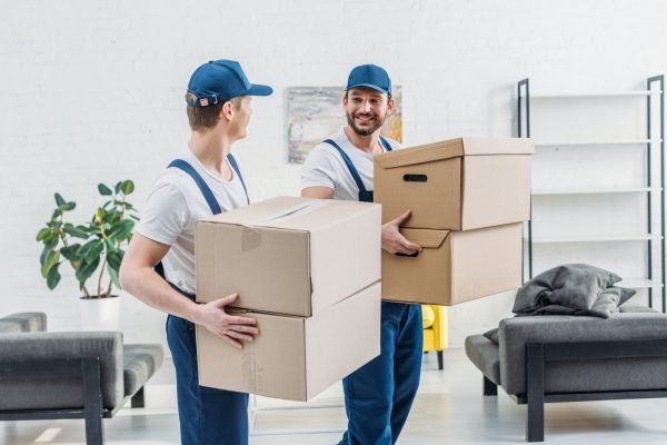 two-movers-in-uniform-looking-at-each-other-while-carrying-cardboard-boxes-in-apartment.jpg